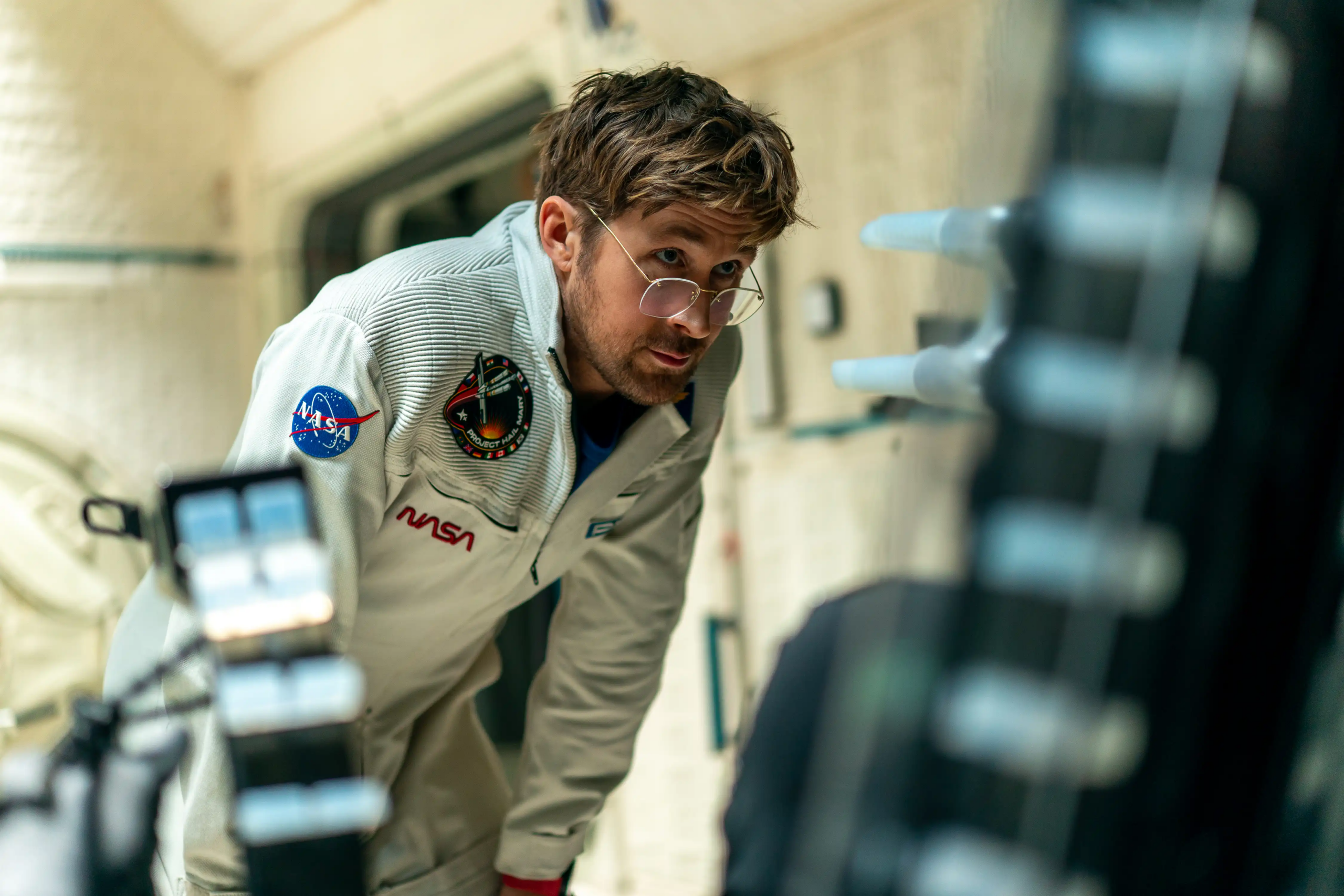 Ryan Gosling as Ryland Grace stares at an experiment while wearing a white NASA uniform.